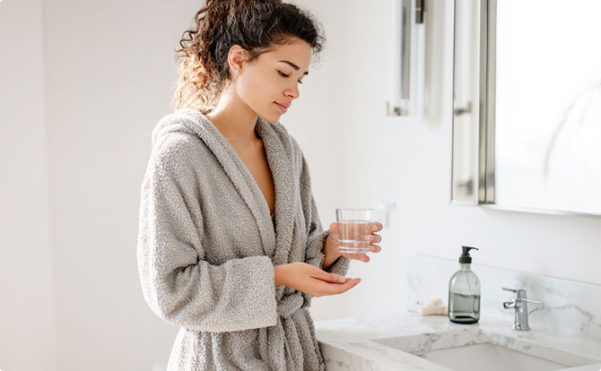 A woman looking at herself in the mirror while taking a white tablet with a glass of water