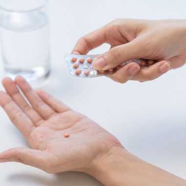 Une jeune femme souriante tenant un verre d’eau et une plaquette de comprimés sur un fond beige.